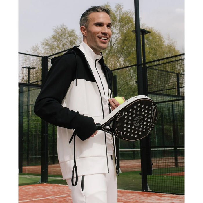 Robin van Persie in a stylish black and white athletic outfit holds a paddle and a tennis ball while smiling on a padel court surrounded by a mesh fence and greenery.