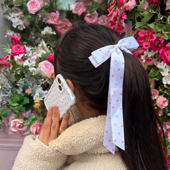 Woman with a ponytail tied with a Coconut Lane mini hearts ribbon bow hair clip, holding a phone among pink flowers.