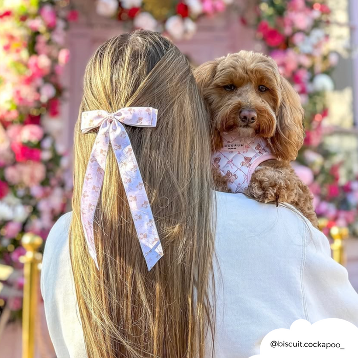 Woman with long hair wears a beige Coconut Lane Thea Teddy ribbon bow clip, holding a brown dog by a flower wall.