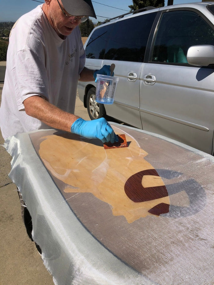 Man using six oz fiberglass cloth on surfboard with resin. Fiberglass is becoming transparent with resin showing below design.