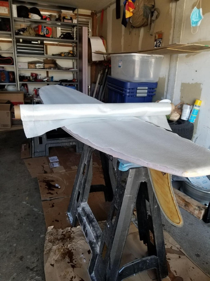 Surfboard blank prepped with fiberglass cloth and roll of fiberglass setting on top. Fiberglass is cut and ready for resin.