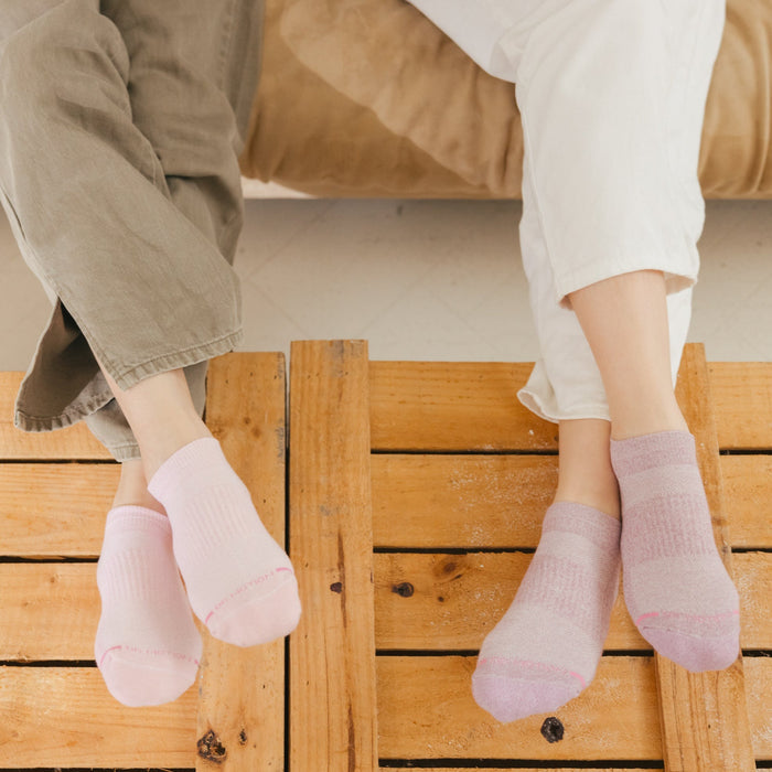 Two pairs of ankle compression socks in pink and light purple, worn by individuals sitting on a wooden surface.