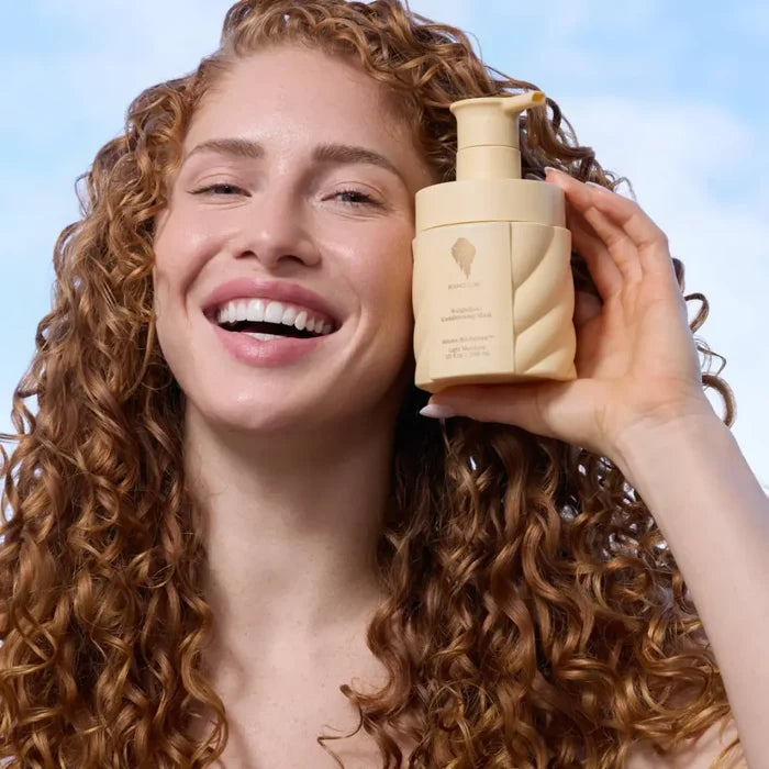 Smiling woman with curly hair holding Bounce Curl conditioner mask for defined, hydrated curls