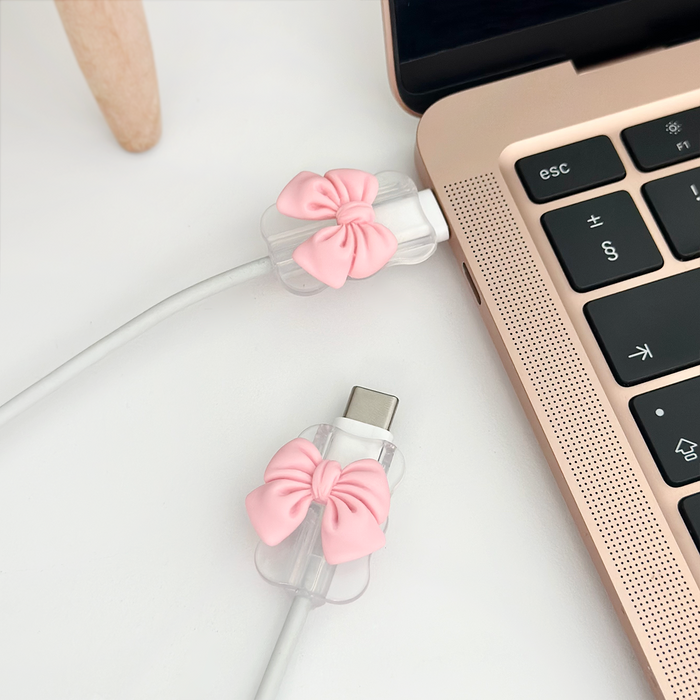 Pink bow cable cover charm from Coconut Lane on a rose gold laptop, clipped around a white USB-C cable.