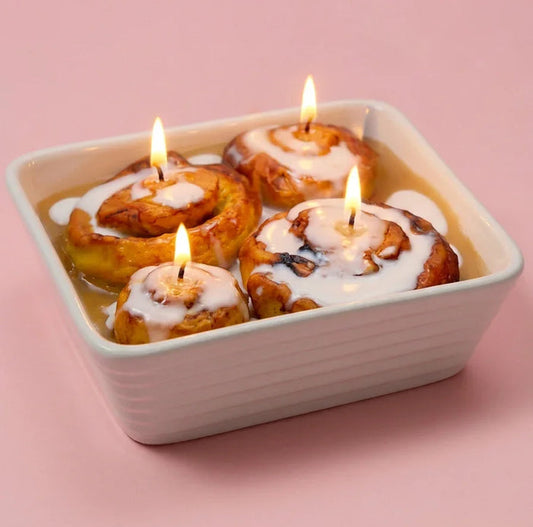 Four cinnamon roll candles with icing in a white dish on a pink background, all lit