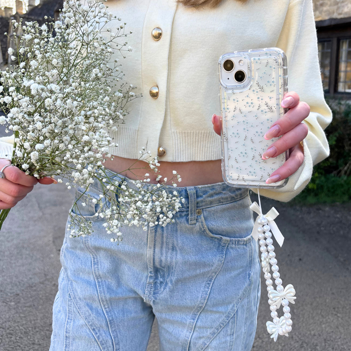 Person in cream cardigan and light blue jeans holds white flowers and a phone with Coconut Lane’s blue floral case.