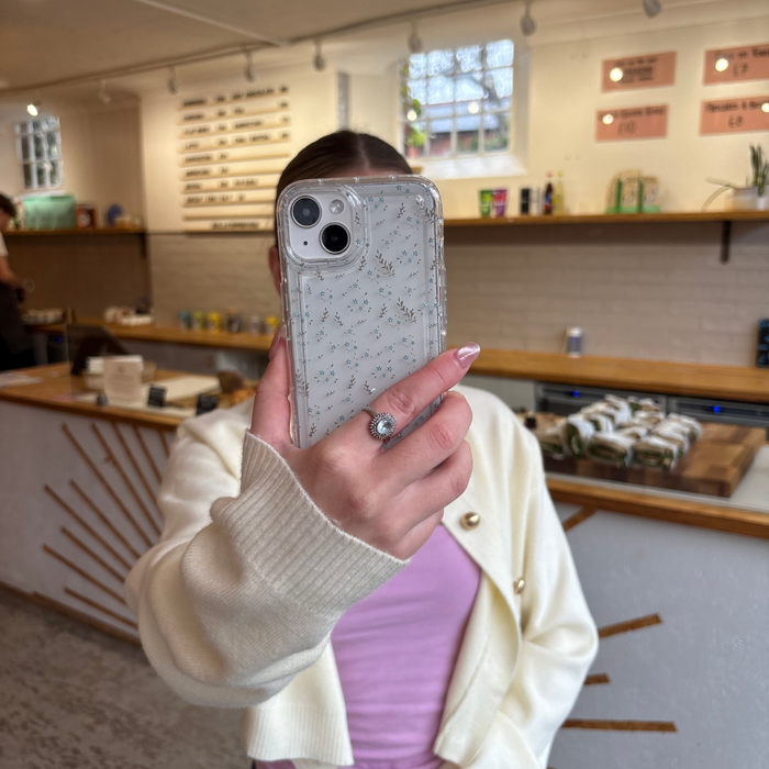 Person in cream cardigan holding Coconut Lane clear phone case with blue floral design at a modern café.
