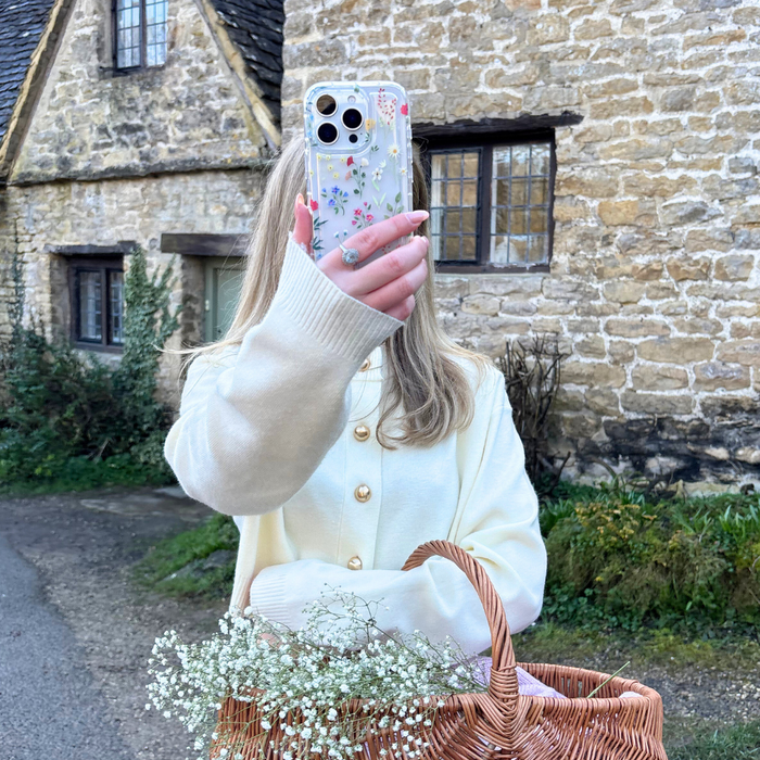 Person in cream sweater holds a clear phone case with colorful wildflower pattern next to a stone house.