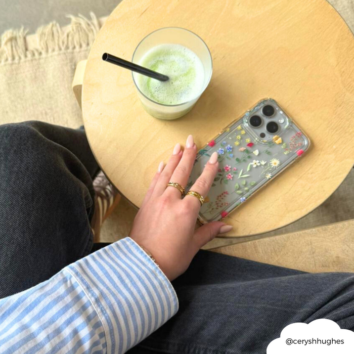 Hand with rings holding a clear phone case with colorful wildflower pattern, next to crossed legs in blue jeans.