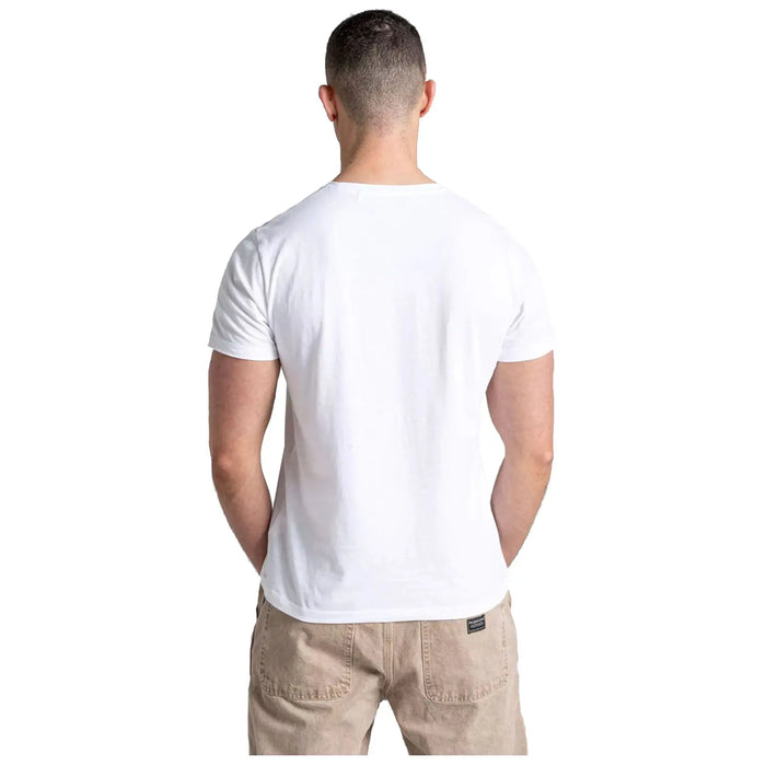 A man with short hair stands facing away wearing a plain white t-shirt and beige trousers in a neutral background creating a simple casual appearance