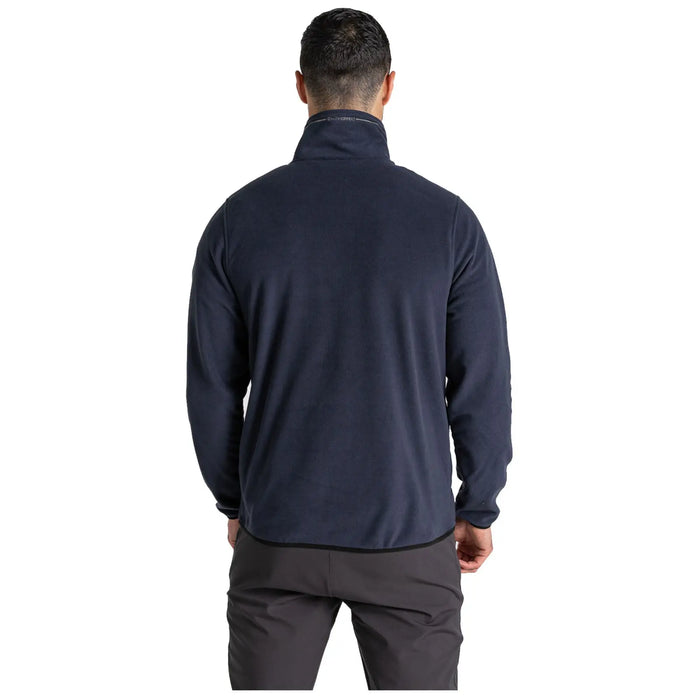A man is standing with his back facing the viewer wearing a dark blue fleece jacket and dark pants in a neutral background suggesting an indoor setting.