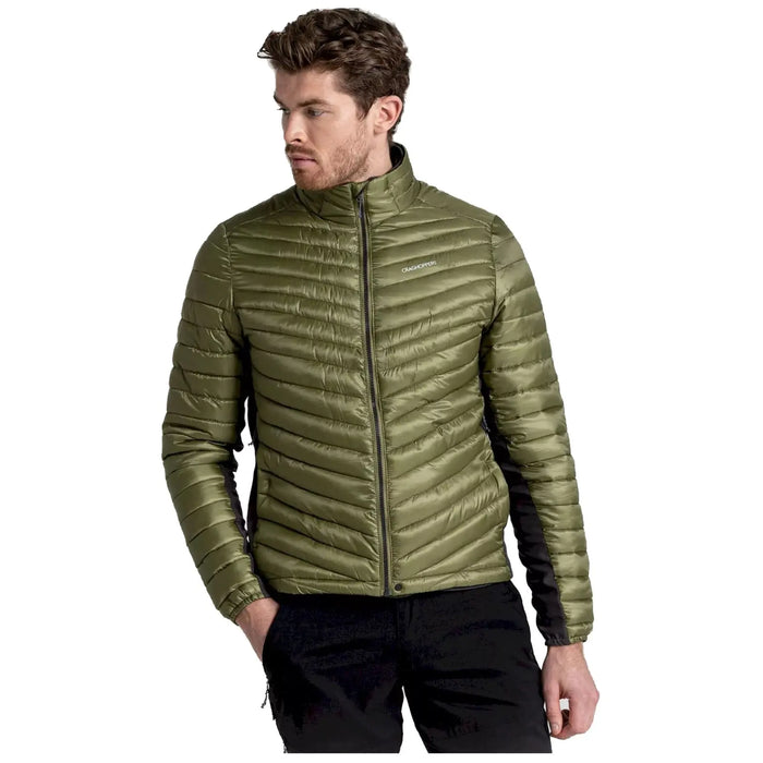 A man wearing a green puffer jacket stands with a serious expression his right hand rested in his pocket while he gazes slightly to his left against a plain background.