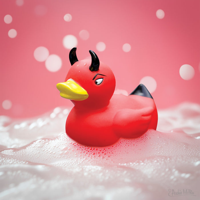 A mischievous Devil Duckie with black horns floats in bubbly water against a pink backdrop decorated with whimsical white bokeh.