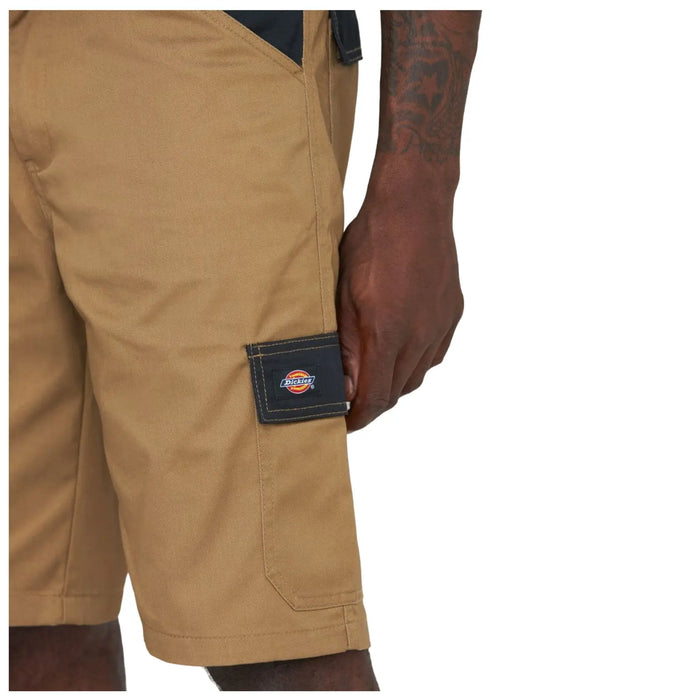 Tan work shorts sit above the knee with a black patch on the side pocket displaying a logo while a hand rests casually on the thigh area in a neutral setting.