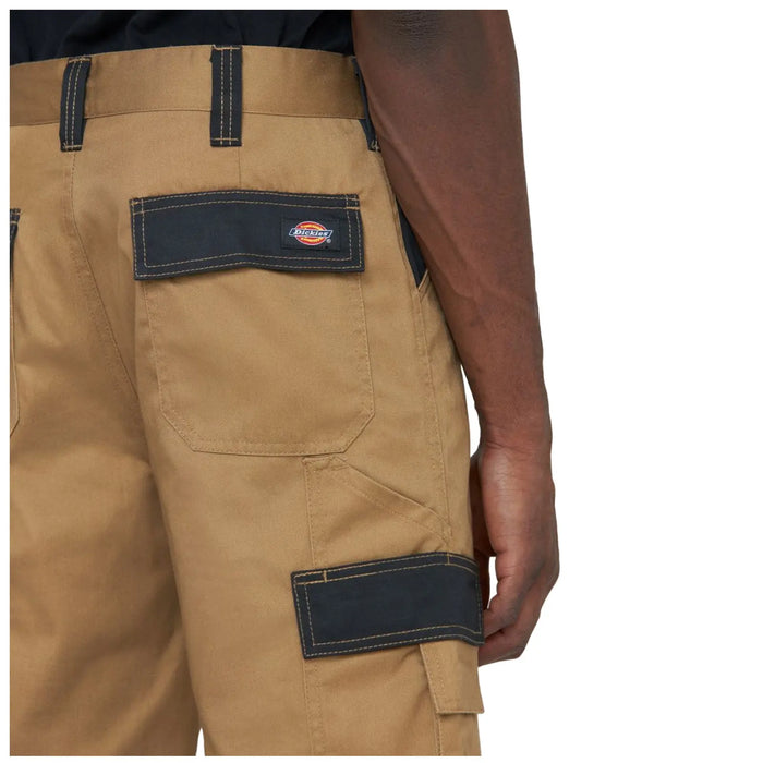 Khaki cargo pants are shown with black accents and pockets The person wearing them is holding the waistband with one hand while their other arm is relaxed by their side