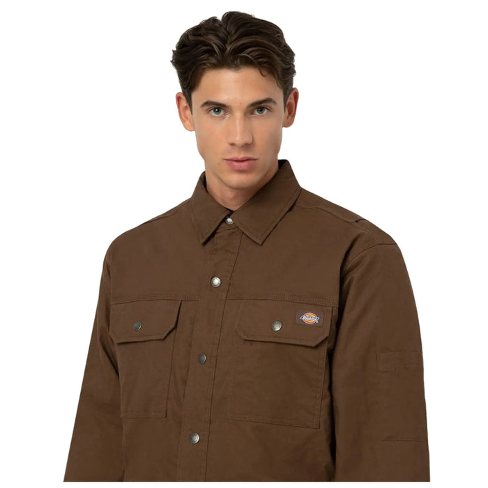 A young man is wearing a brown jacket with a collar and two front pockets, standing against a plain background, looking directly at the viewer with a serious expression.