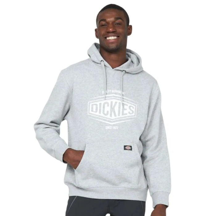 A young man is smiling while wearing a gray hoodie with a logo on the front. He has one hand in the pocket and is standing against a plain background.