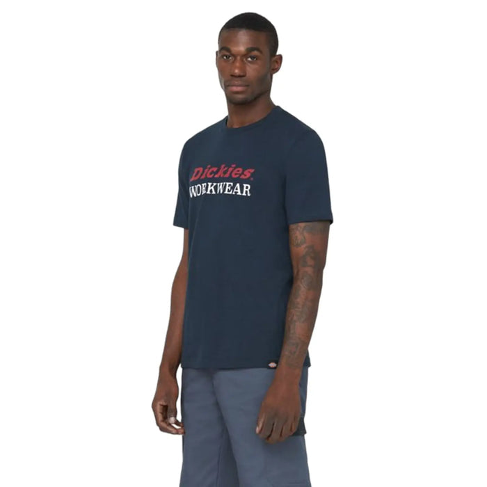 A man stands confidently wearing a dark blue t-shirt with "Dickies WORKWEAR" printed in red and white, paired with gray shorts in a light background.