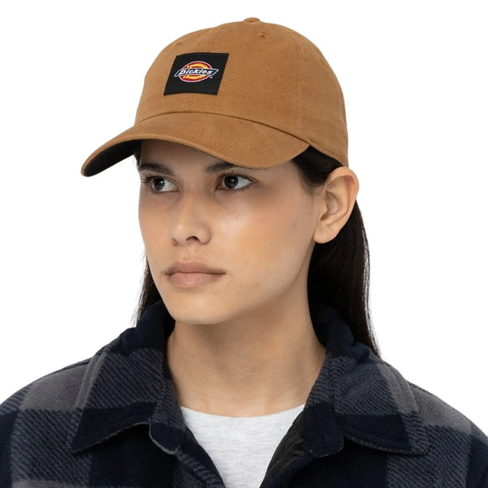 A person wears a brown cap featuring a logo while looking slightly to the side. They are dressed in a black and gray plaid shirt, standing against a plain white background.