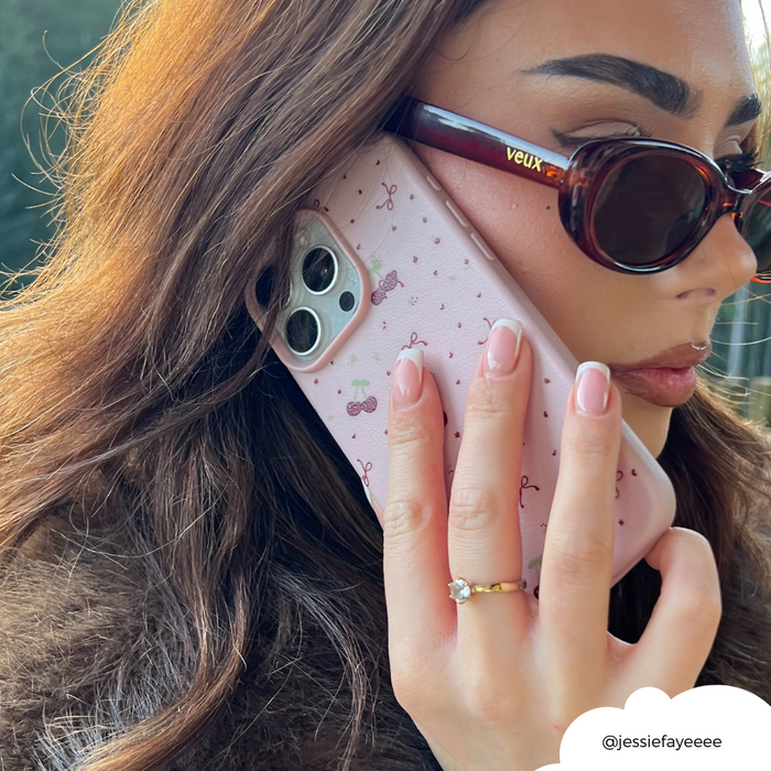 Woman with sunglasses holds a pink phone case with bows and cherry pattern, showing her ring and manicured nails.