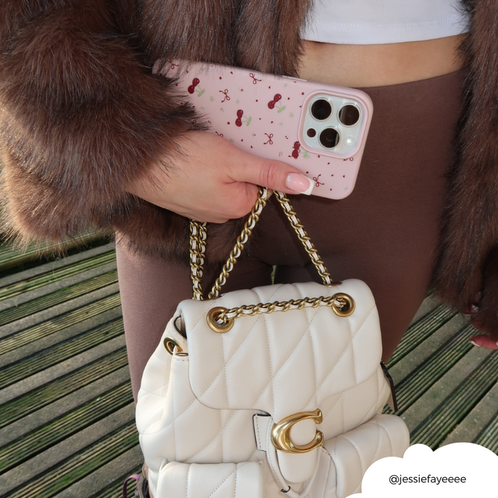 Person in brown pants and faux fur coat holding a white handbag and a pink phone case with bows and cherries pattern.