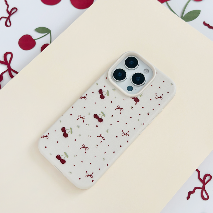 Soft white phone case with bows and cherry pattern, shown on a cream surface with scattered cherry accents.