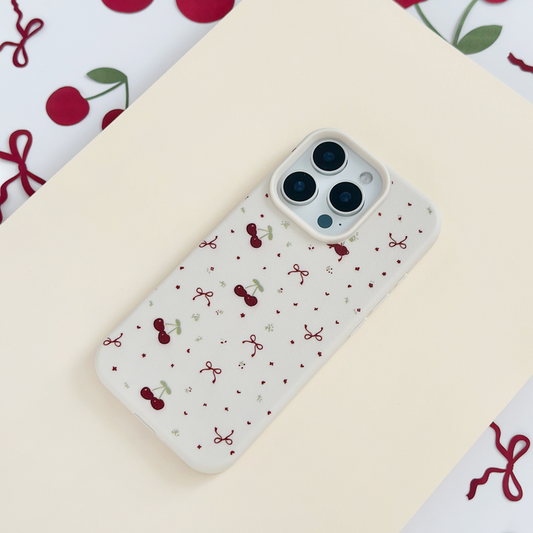 Soft white phone case with bows and cherry pattern, shown on a cream surface with scattered cherry accents.
