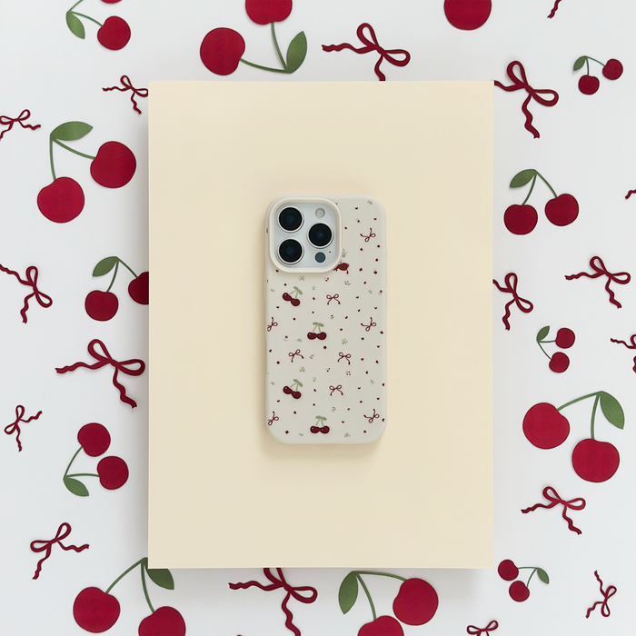 Soft white phone case with red cherry and pink bow pattern, displayed on cream paper.