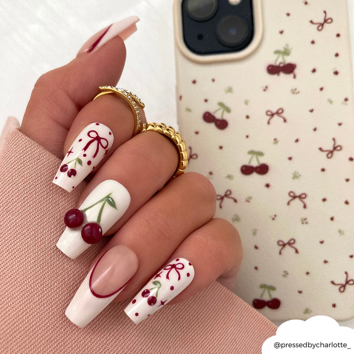 White coffin nails with red cherry and bow designs beside a soft white phone case with matching cherry and bow pattern.