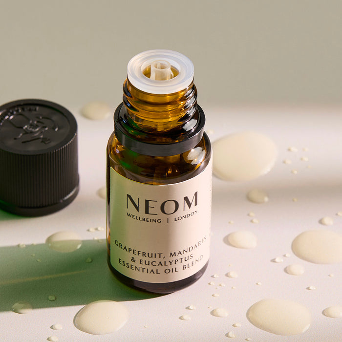 Open amber glass bottle of NEOM Grapefruit, Mandarin & Eucalyptus Essential Oil Blend on a white surface with droplets of oil around it.