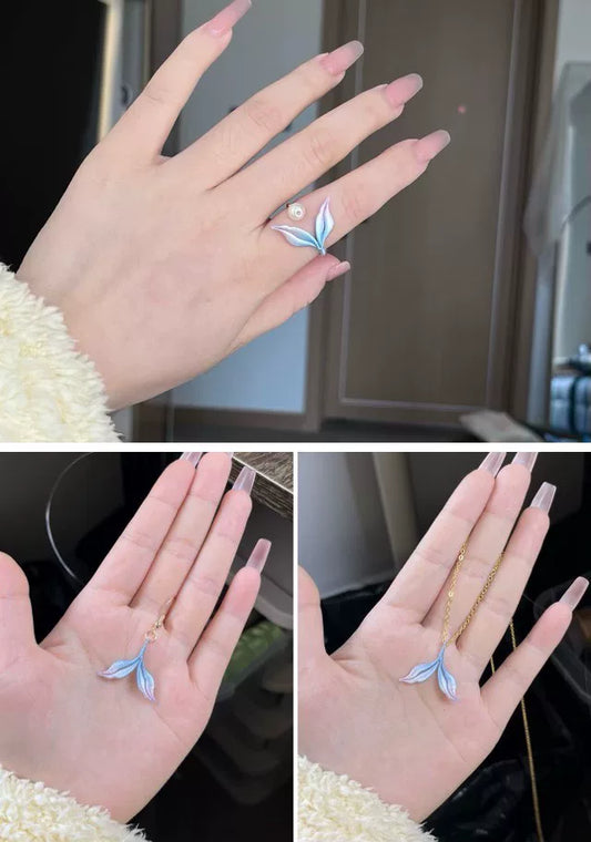 A hand wearing a blue mermaid tail ring and holding a blue mermaid tail necklace