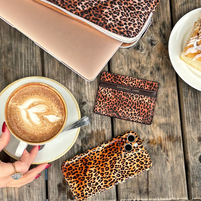 Hand holding a latte beside a laptop, pastry, and Coconut Lane leopard print card holder on a wooden table.