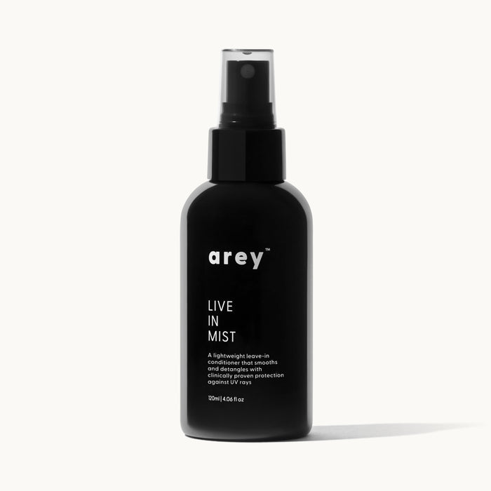 Product shot of arey Live In Mist dark glass spray bottle on a white/cream background. Label reads: arey™ LIVE IN MIST — "A lightweight leave-in conditioner that smooths and detangles with clinically proven protection against UV rays." 120ml | 4.06 fl oz.