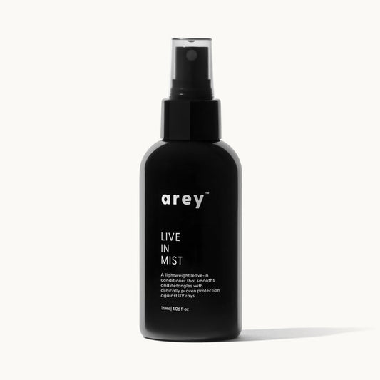 Product shot of arey Live In Mist dark glass spray bottle on a white/cream background. Label reads: arey™ LIVE IN MIST — "A lightweight leave-in conditioner that smooths and detangles with clinically proven protection against UV rays." 120ml | 4.06 fl oz.