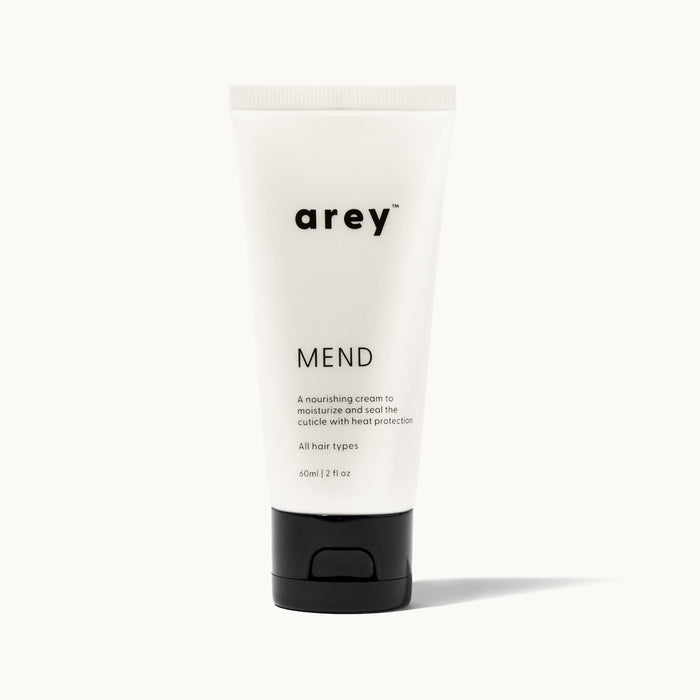 Product shot of Arey Mend hair treatment tube on a white/cream background. Label reads: arey® Mend. "A nourishing cream to moisturize and seal the cuticle with heat protection". All hair types. 60ml/2 fl oz.