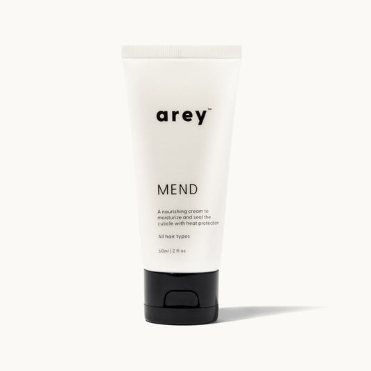 Product shot of Arey Mend hair treatment tube on a white/cream background. Label reads: arey® Mend. "A nourishing cream to moisturize and seal the cuticle with heat protection". All hair types. 60ml/2 fl oz.