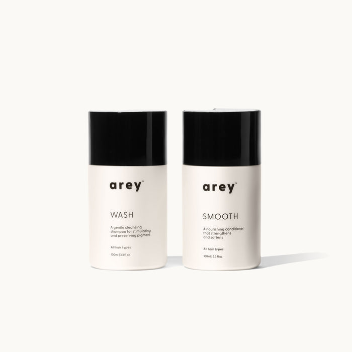 Two Arey Mini Duo travel-size bottles side by side on a white background. Left: arey® Wash — "A gentle cleansing shampoo for stimulating and preserving pigment", All hair types, 100ml/3.3 fl oz. Right: arey® Smooth — "A nourishing conditioner that strengthens and softens", All hair types, 100ml/3.3 fl oz.