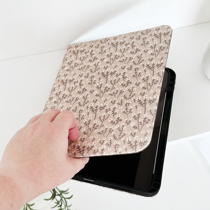 Hand opening a Coconut Lane iPad case with nude flower pattern on a white surface, small plant beside it.