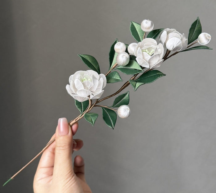 Handmade white Pearl Jasmine Chanhua flower branch with green leaves and buds, held by a hand against a grey background, representing a DIY craft floral arrangement.