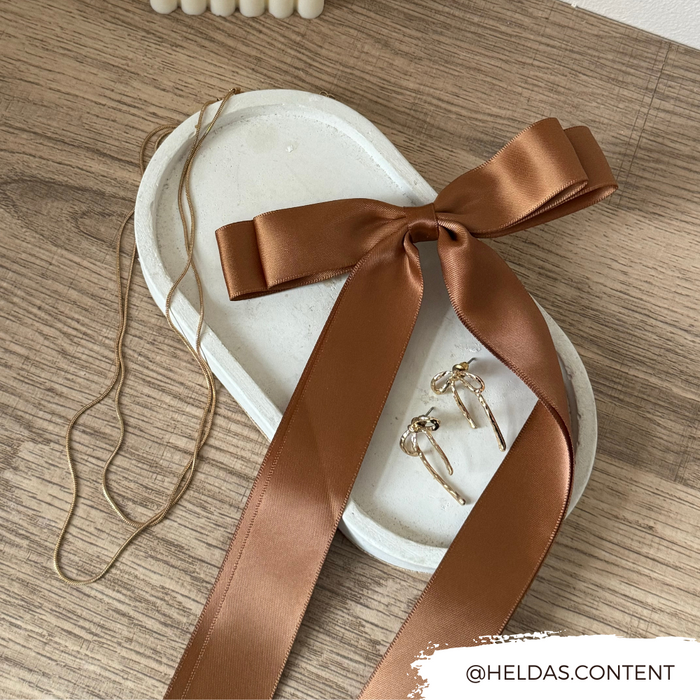 Brown ribbon bow hair clip with gold necklace and earrings displayed on a white tray on a wooden surface.