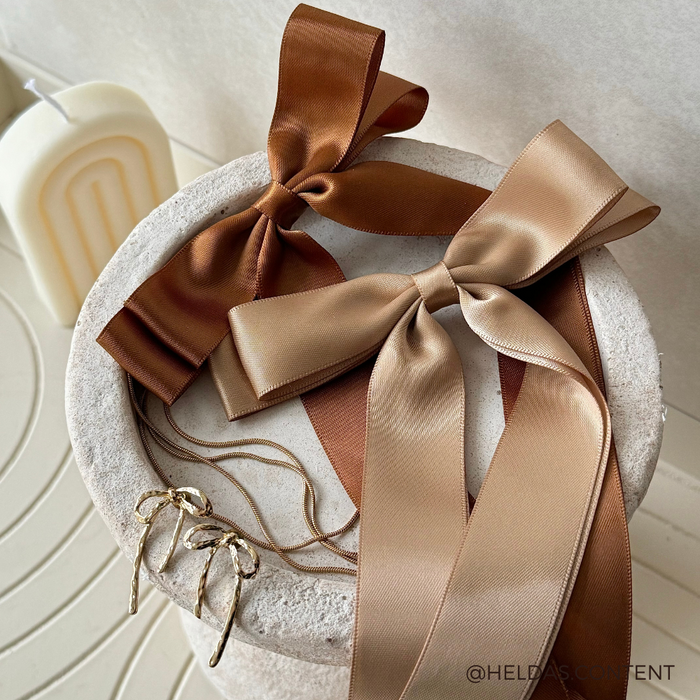 Brown ribbon bow hair clips and gold hoop earrings displayed on a textured round pedestal.