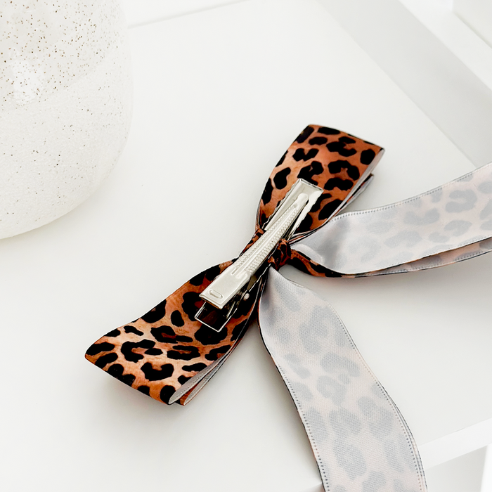Leopard print ribbon bow hair clip with metal clasp, displayed on a white surface beside a speckled vase.