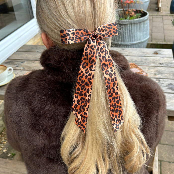 Woman with long blonde hair wearing a Coconut Lane leopard-print ribbon bow clip, seated outdoors in a fur coat.
