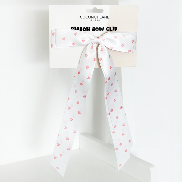 White satin ribbon bow clip with pink hearts pattern, attached to branded Coconut Lane card on a white background.