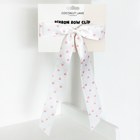 White satin ribbon bow clip with pink hearts pattern, attached to branded Coconut Lane card on a white background.