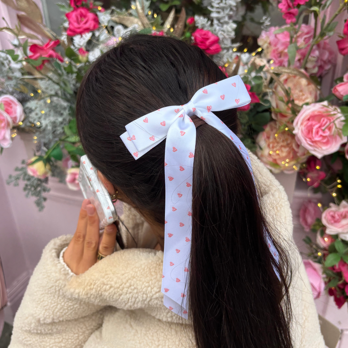 Woman with long dark hair wears a Coconut Lane mini heart-patterned ribbon bow clip, surrounded by pink flowers.