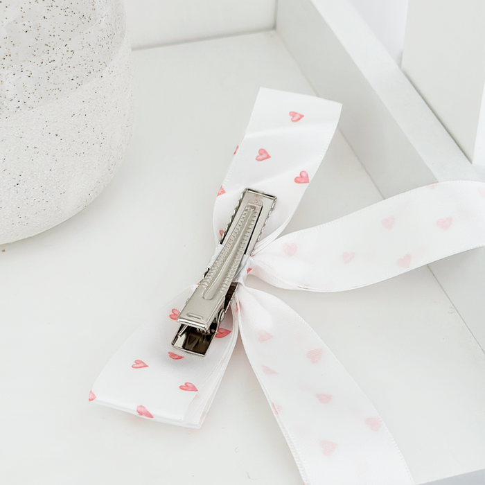 White ribbon bow clip with pink hearts, placed on a white surface beside a speckled container.