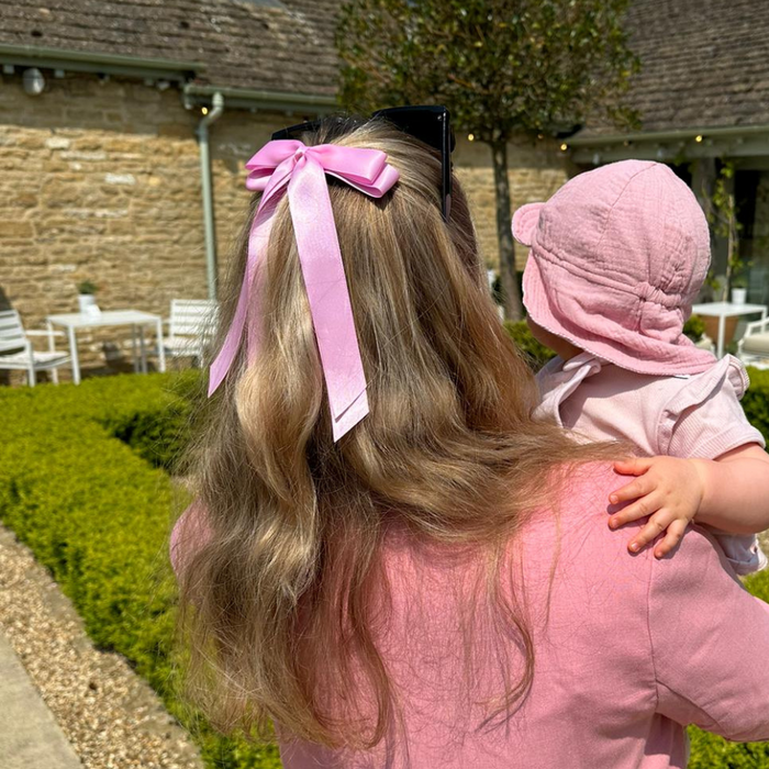Woman wears a pink Coconut Lane ribbon bow hair clip, holding a baby in a pink hat outdoors near a stone building.