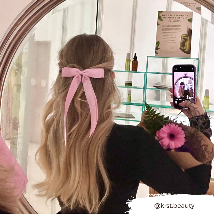 Woman with long blonde hair wears a Peony Pink ribbon bow clip from Coconut Lane, holding flowers in a mirror selfie.