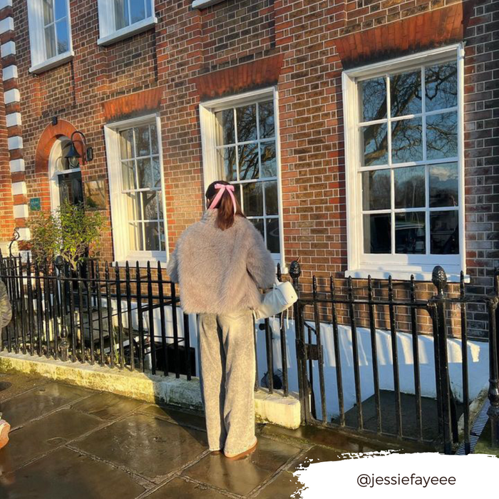 Woman in a furry jacket wears a pink Coconut Lane ribbon bow clip, standing by a brick building on a sunny day.